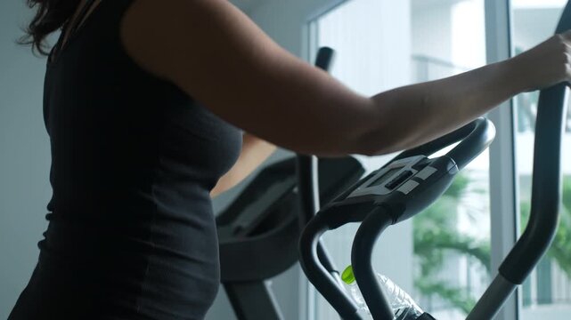 Young woman working out on an elliptical machine in a modern gym pausing mid-session to drink water from a bottle before continuing her exercise. Fitness concept