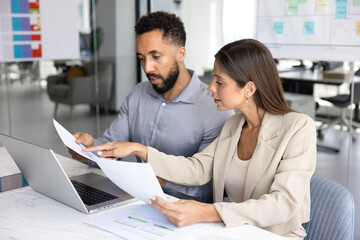 Fototapeta premium Two business professionals, man and woman colleagues collaborating in modern office seated at desk, reviewing printed reports and data while using laptop, focused on teamwork and strategic planning