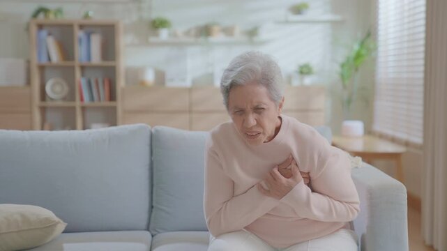 elderly woman experiences chest pain and acute heart failure on the couch due to a heart attack from cardiac arrest, older or patient with difficulty breathing due to vascular. medical emergency