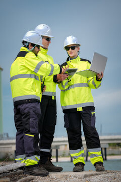 Construction engineer safety hardhat high visibility jacket laptop tablet team discuss project plan during outdoor site inspection near industrial harbor, focused collaboration