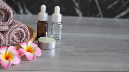 Luxury spa still life with essential oil dropper bottles (amber and clear glass), a scented candle, soft pink mauve towels tied with rope, and pink frangipani flowers on a light gray wooden table. Pre © kanjana