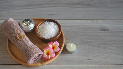 Minimalist spa still life with white bath salt in a wooden bowl, pink frangipani flowers, mauve rolled towel, and a scented candle on a light gray wooden table. Soft focus background with extensive co © kanjana