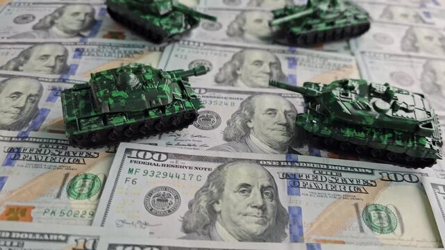 Small tanks move on top of dollar bills in a playful setup with a mix of colors and textures