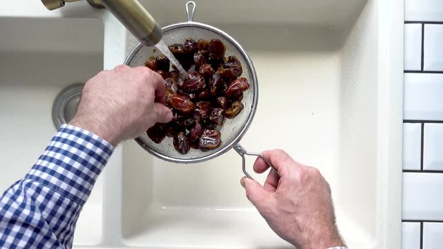 Male hands wash dried dates in a mesh strainer under running water in a kitchen sink. Concept of healthy eating and using natural sweeteners. Top view. Ideal for nutrition, vegan diet, cooking content