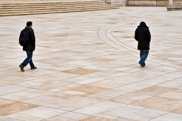Two individuals walking separately across an expansive open paved plaza on a cold day in winter