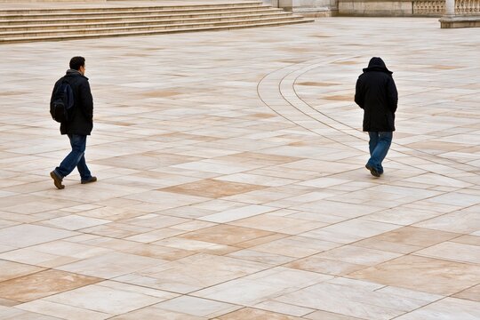 Two individuals walking separately across an expansive open paved plaza on a cold day in winter