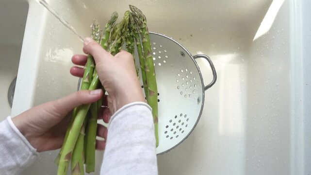Female hands wash asparagus under running water over a metal colander in a kitchen sink. Concept of healthy eating and using natural sweeteners. Ideal for nutrition, vegan diet, and cooking content
