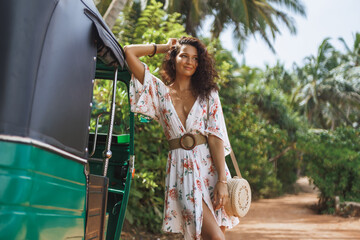 Obraz premium Woman in floral dress standing by green tuk-tuk on tropical dirt road