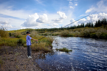 Naklejka premium A young boy stands on the rocky bank of a river while fishing with a long rod under a dramatic blue sky filled with white clouds. He enjoys a peaceful outdoor adventure in nature.