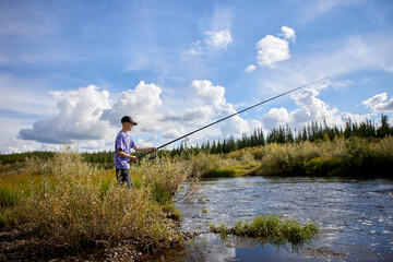 Naklejka premium A teenage boy fishing with a long rod on a scenic riverbank under a dramatic blue sky. Surrounded by wild grasses and forest, he enjoys a peaceful outdoor recreation moment in untouched nature.