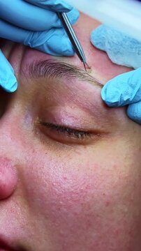 A woman is lying down while a beautician works on her eyebrows. The beautician is holding tweezers and focusing on shaping the brows carefully.