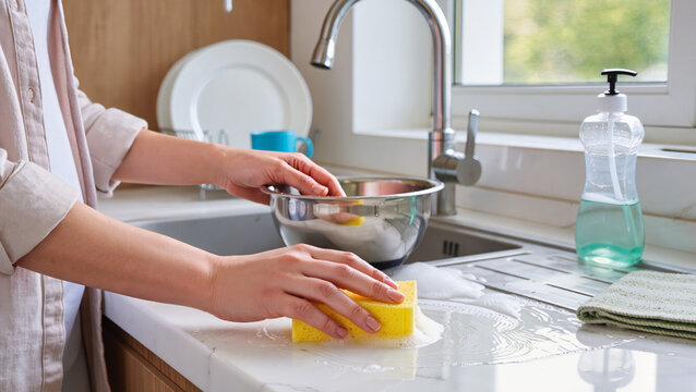 Wiping kitchen surfaces, Washing dishes in the sink, Kitchen cleaning routine, Housekeeping and countertop maintenance, Disinfecting kitchen sink area, Using sponge for cleaning