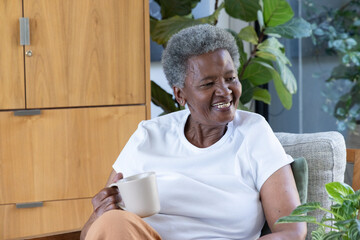 Senior African American woman sitting in armchair at home holding ribbed mug near potted plants © wavebreak3