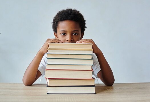 Young Black boy peeking over a tall stack of books. Education and literacy concept. African American child student with pile of books on a wooden table. Copy space for text