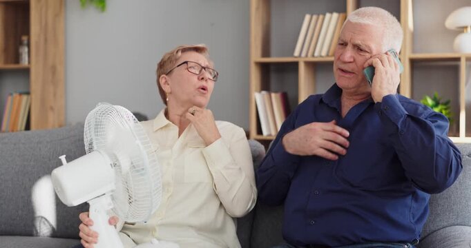 Seniors couple in heatwave overheating phone fan repair. At home in summer they look in distress as man calls technician about a broken air conditioner. Concept highlights home cooling system failure.