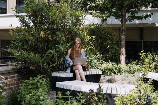Woman sits on bench in courtyard garden working on laptop computer. Academic digital campus, outdoor research study, online course assignment, university students learning.