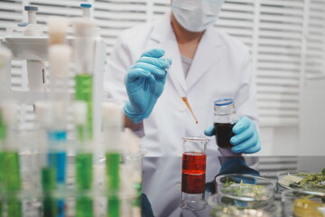 Fototapeta premium Scientist in mask and gloves testing plant extract with a dropper in a laboratory, with glassware, herbs, and red liquid on the workbench.
