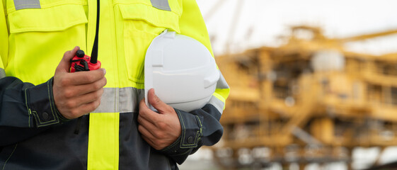 Fototapeta premium Industrial engineer in a high-visibility safety jacket and earmuffs, holding a hard hat and walkie-talkie radio at an offshore energy platform facility. Concept of workplace safety