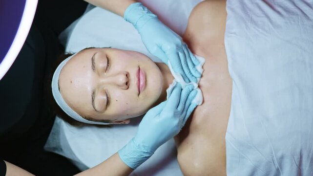 A woman lies on a treatment table while a professional applies a facial mask. The setting has soft lighting and a clean, organized appearance. The woman seems relaxed and comfortable.