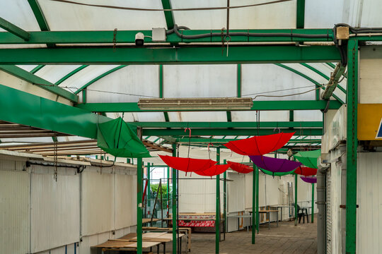 Interior of an empty marketplace under a translucent roof with a green metal structure, featuring open colorful umbrellas in red, purple, and green hanging down, surrounded by closed white market stal