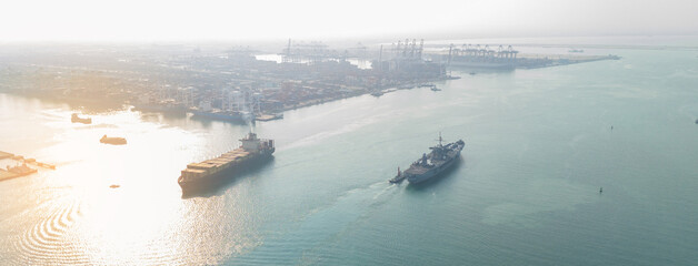 naval warship escorting a large commercial container ship near a busy industrial port, symbolizing maritime security, naval patrol, global trade protection, and geopolitical stability. © Yellow Boat