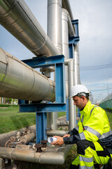Naklejka premium industrial engineer wearing a white hard hat and high-visibility PPE squats to point at a large metal pipeline while speaking into a walkie-talkie during an outdoor refinery inspection.