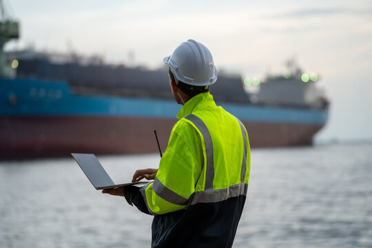 Marine engineer using a laptop to monitor fleet operations with a blurred container ship in the background. Concept of smart port management, digital maritime logistics, and supply 