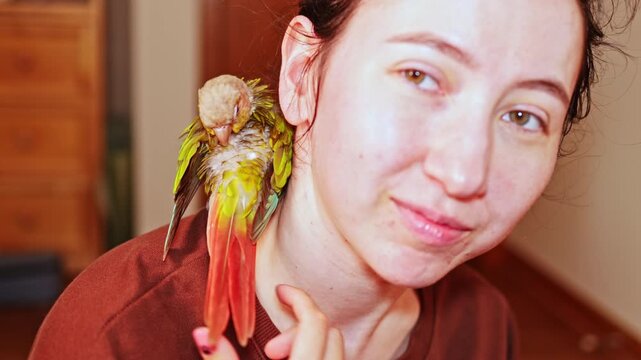 A woman is indoors with a green and red parrot perched on her shoulder. They share a light moment as she smiles gently. It is afternoon and the setting is cozy.