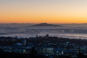 Fototapeta premium Auckland, New Zealand. Rangitoto Island emerges from the morning fog as the sun rises over the Hauraki Gulf, casting a warm glow on the city skyline.