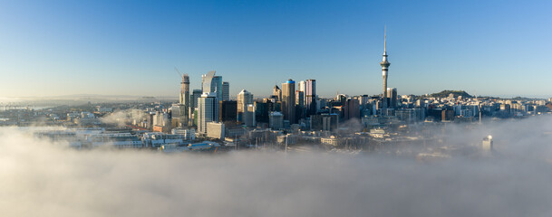 Obraz premium A stunning aerial view of Auckland, New Zealand, as low-lying fog blankets the harbor and city center, revealing the iconic Sky Tower and skyscrapers on a clear morning.