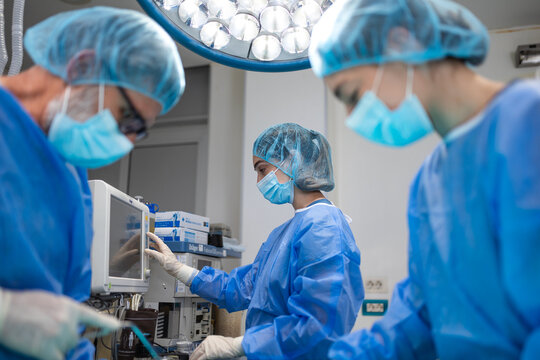 Medical team in blue scrubs and surgical masks monitors patient vitals on a digital screen in a modern hospital operating room during a surgical procedure.