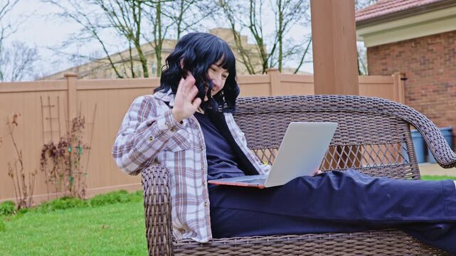 A young person sits on a wicker sofa in a backyard, waving at the camera while using a laptop. Trees and a fence are seen in the background during cloudy weather.