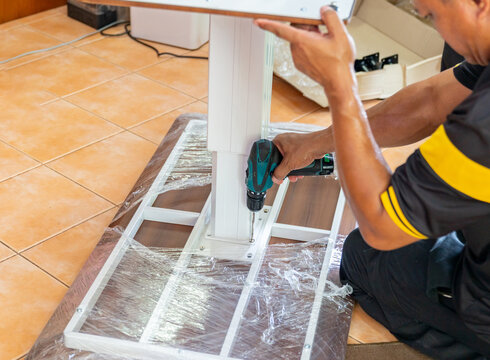 Professional technician using a cordless electric drill to screw and assemble a height adjustable desk frame onto a wooden tabletop inside a house.