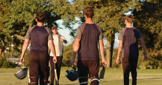 Diverse male cricket players approaching coach on sunlit field, tapping bats, carrying helmets
