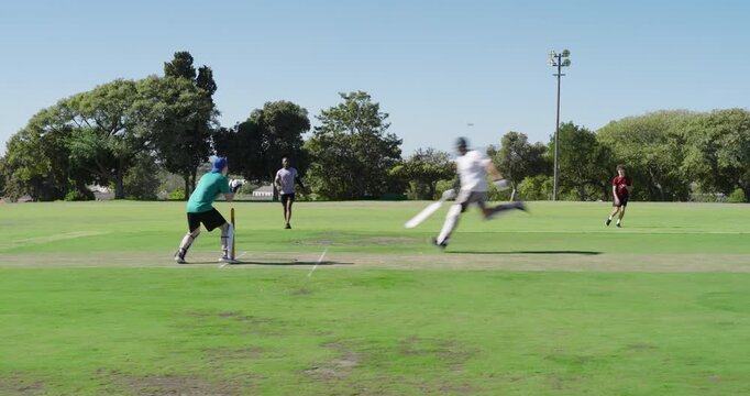 Asian wicket keeper adjusting on pitch as bowler sprinting, fielder hurling ball
