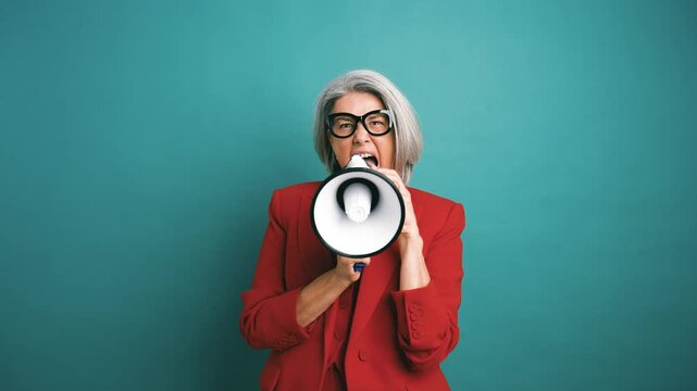 Confident senior woman in a red suit energetically shouting and protesting with a megaphone. Female leader making an announcement against a solid background