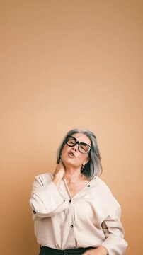 Mature stylish woman with grey hair and glasses massaging her sore neck. Exhausted lady feeling tired due to neck pain and muscle tension. Vertical