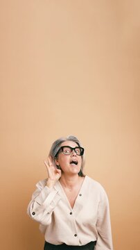 Expressive elderly woman with grey hair and glasses cupping her ear, trying to overhear a secret conversation and gossiping. Vertical
