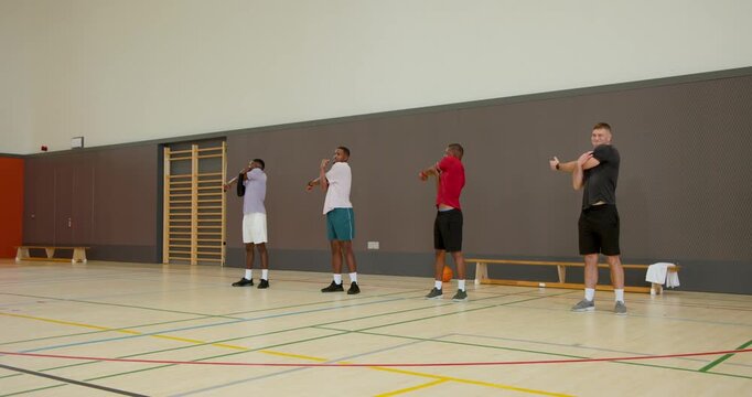 Diverse male teammates stretching arms wearing sportswear on gym court, with basketball and towel