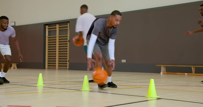 Dribbling man in black shorts weaving orange basketball around training cones on gym court