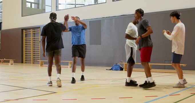 Orange ball rolling toward diverse male teammates at gym, intercepting, celebrating high-fives