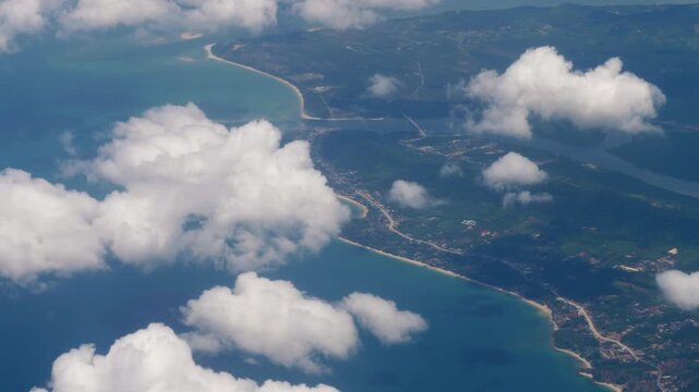 Aerial view of white clouds floating over tropical island coastline and turquoise ocean, shot from high altitude airplane window during day.