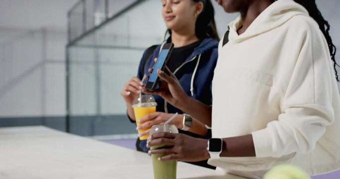 Diverse female friends placing phone on juice and smoothie cups, sharing workout progress at court