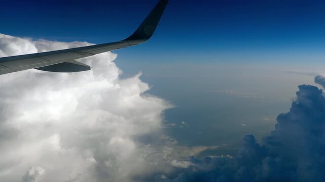 Airplane wing flying over massive fluffy white clouds and blue ocean under bright daylight, wide angle passenger window POV