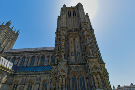 A dramatic, low-angle view of Wells Cathedral's majestic stone tower. Ornate Gothic architecture rises against a bright, sunny blue sky