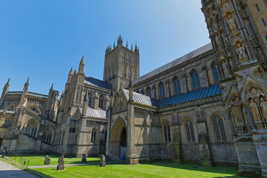 A grand stone cathedral with ornate Gothic architecture and a tall central tower stands under a clear blue sky - Wells Cathedral - Wells - Somerset - UK