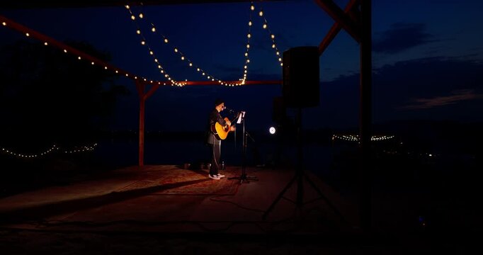 Solo musician playing acoustic guitar under lit pergola on a dusk beach