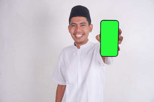 Muslim Asian man showing smartphone green screen with laughing, smiling, shocked and excited expressions. Muslim man in muslim costume, pecis standing smiling and pointing to blank mobile phone.