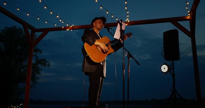 Male musician playing acoustic guitar on lakeside wooden stage at dusk