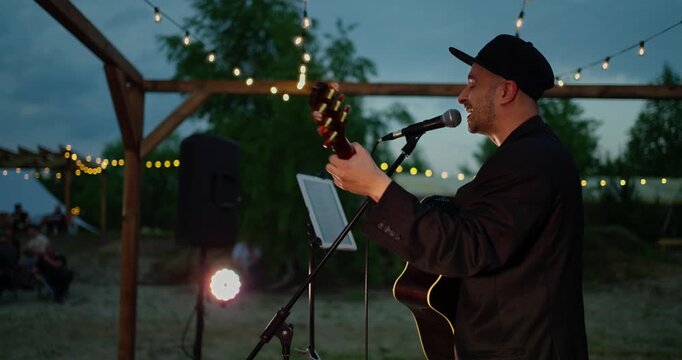Male acoustic guitarist performing under string lights at outdoor evening pergola